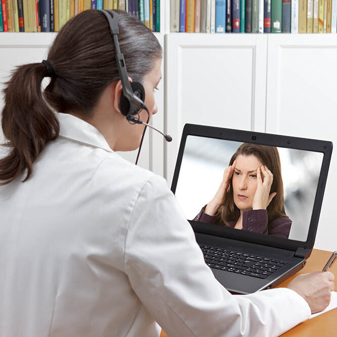 Doctor sitting at the desk of her office with headset and laptop, making a video call with a patient.